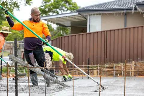 Concreters in hi-vis and wide-brim hats screeding a new slab beside a Colorbond fence
